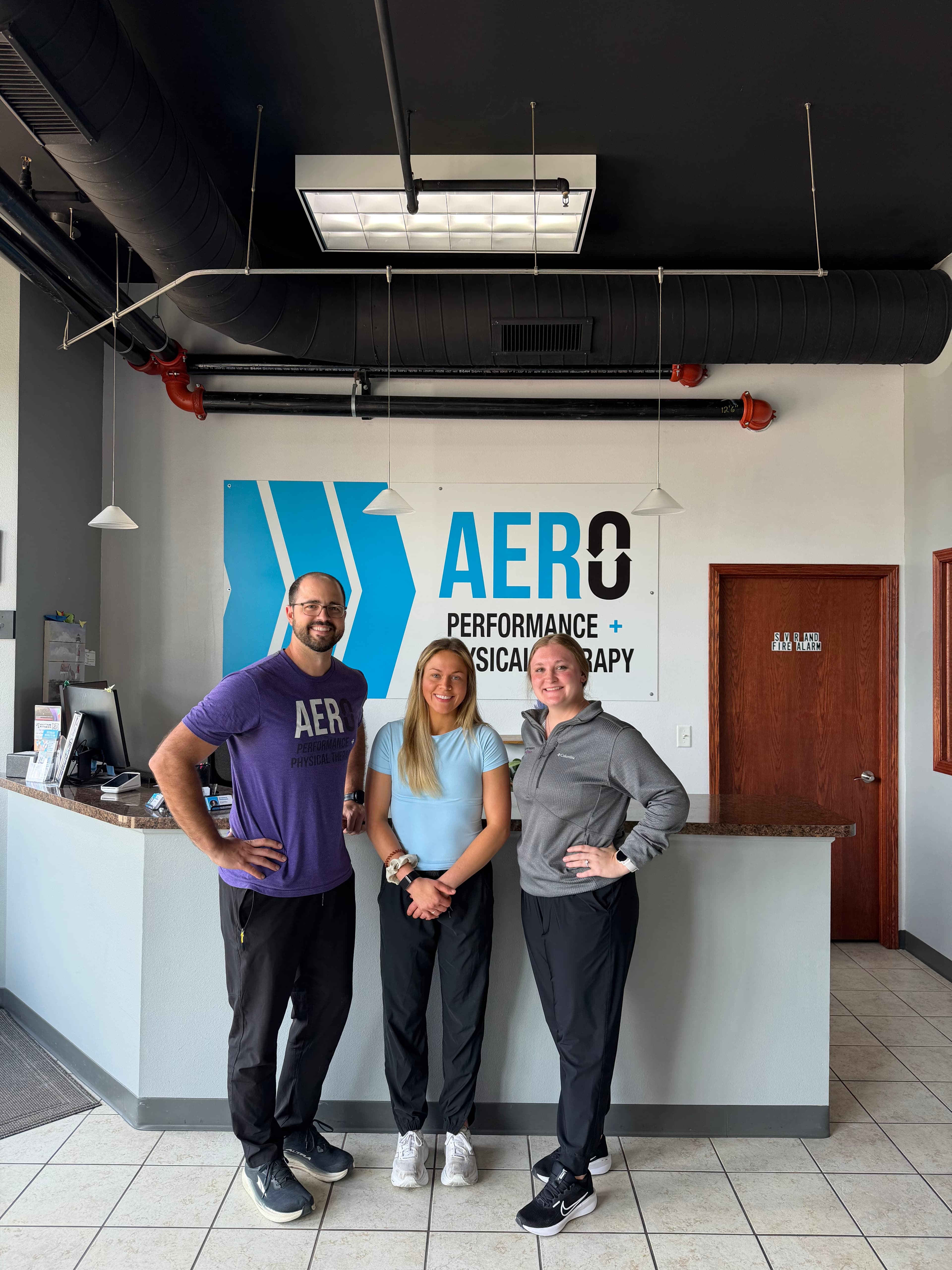 The Aero Performance and Physical Therapy team standing together inside the clinic's lobby in Coralville, Iowa, in front of the Aero logo wall