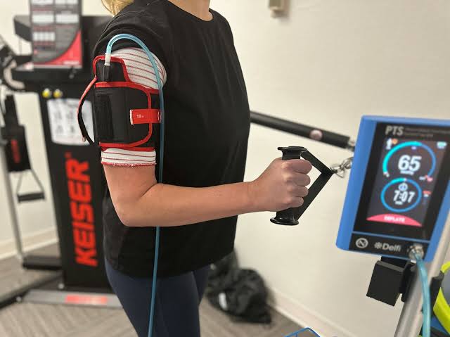 Patient wearing a personalized blood flow restriction cuff on her upper arm while performing a resisted arm exercise during BFR training at Aero Performance in Coralville, Iowa