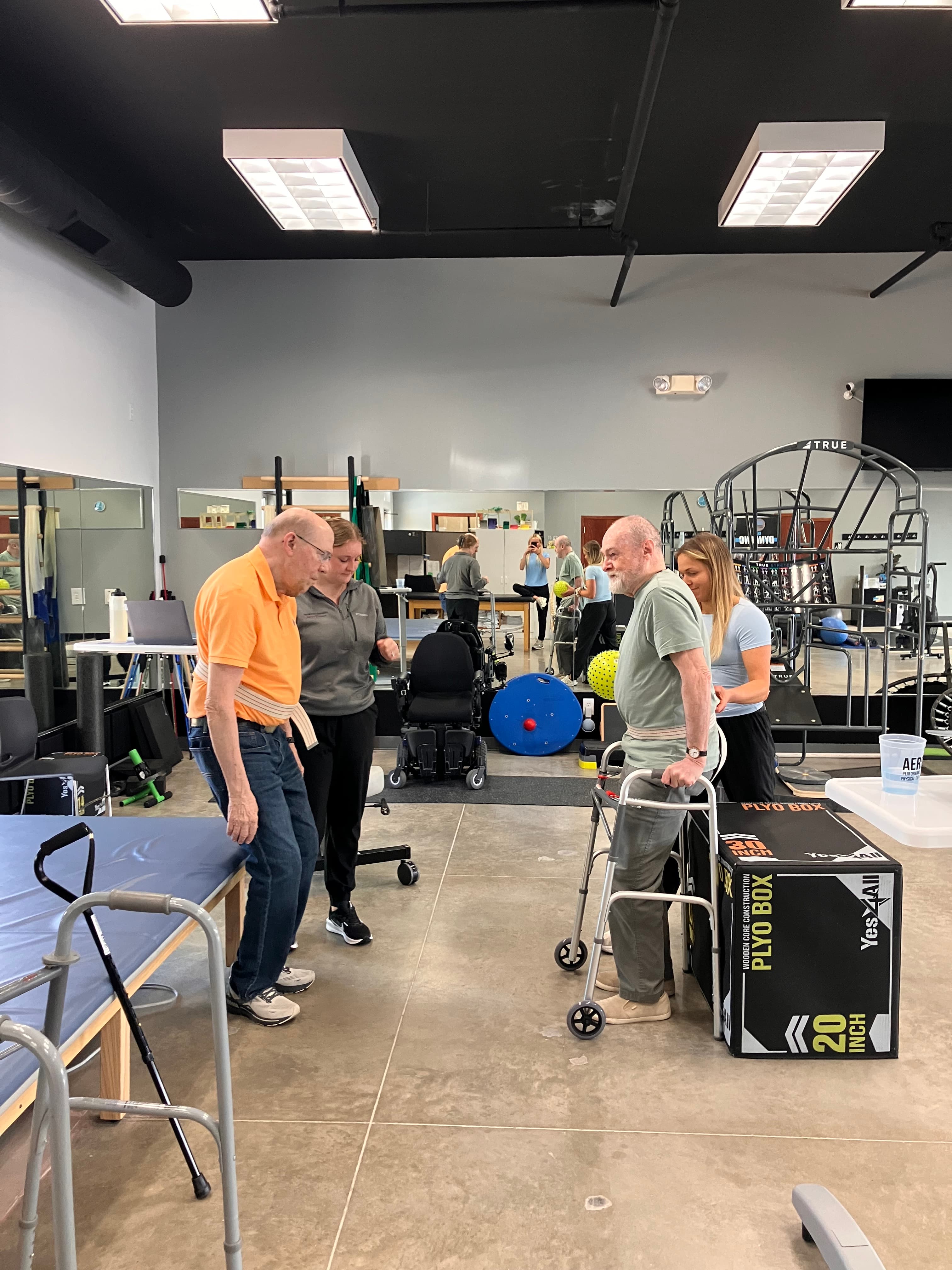 Aero Performance physical therapy staff assisting an older adult patient with gait and strength training using parallel support bars in the clinic's mobility gym