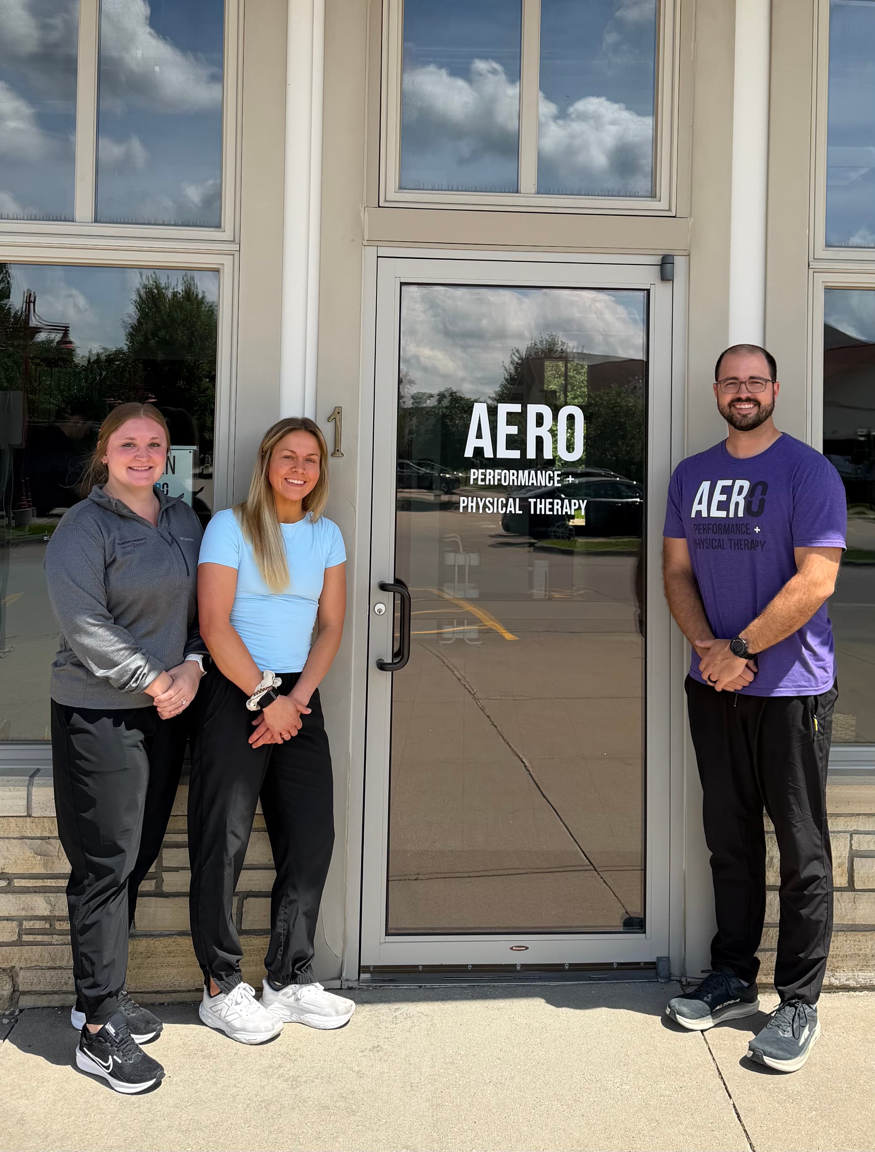 Three Aero Performance and Physical Therapy team members standing together outside the clinic's front entrance in Coralville, Iowa, wearing Aero-branded apparel